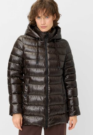 Woman wearing a shiny dark brown quilted puffer jacket with hood and front zipper, standing against a plain light background.