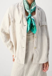 Giacca in lino beige chiaro con due tasche frontali, chiusura a bottoni e vestibilità comoda, abbinata a una sciarpa di seta con motivo turchese.