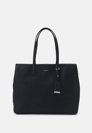 Zwarte Calvin Klein draagtas met dubbele handvatten en een klein bevestigd label tegen een eentonige achtergrond.