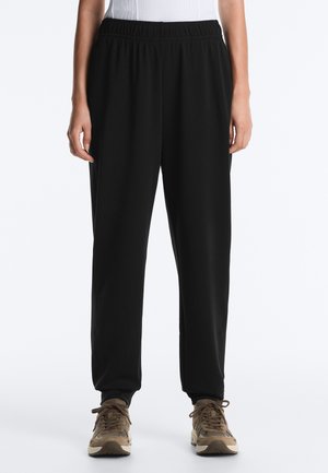 Zwarte losse broek met elastische taille, gedragen met beige sneakers, getoond van taille tot voeten bij een staand persoon.