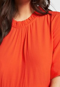 Haut rouge à manches courtes avec un col froncé, présentant une texture lisse et un tissu fluide. La couleur est vive et accrocheuse.