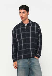 Hombre con una chaqueta negra de botones con líneas de cuadros blancos y jeans azules, de pie con una mano en el bolsillo, mirando hacia un lado.