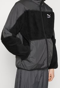 Puma jakke med en blanding af struktureret sort fleece og ternet stof i grå nuancer, med lynlåslukning foran og et diskret logo på brystet.