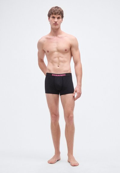 Fekete boxeralsó texturált mintával, rózsaszín márkajelzéses derékpánttal. A modell mezítláb áll, bemutatva a fazont és a kialakítást.