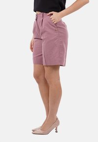 Pantaloni tailored rosa chiaro con pieghe, tasche laterali e chiusura con bottone. Indossati con una maglietta nera e tacchi nudi.