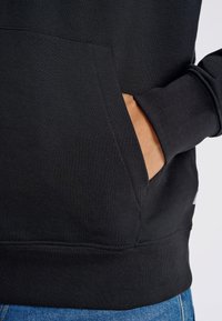 Sudadera negra con un bolsillo tipo canguro, puños y dobladillo acanalados, y una textura suave. Presenta una etiqueta lateral, colocada sobre unos vaqueros azules.