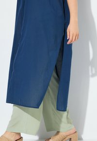 Granatowa, długa tunika z bocznym rozcięciem, w zestawieniu z jasnozielonymi spodniami o szerokich nogawkach. Modelka nosi beżowe, plecione sandały na słupku.