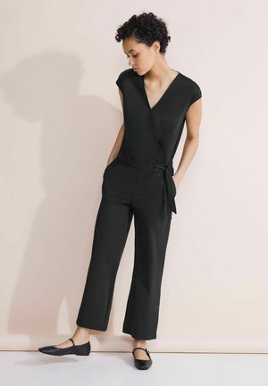 Jumpsuit nero avvolgente con maniche corte, scollatura a V, vita elastica e tasche laterali. Abbinato a scarpe piatte nere su uno sfondo chiaro.
