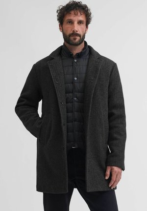 Férfi, aki sötétszürke gyapjú kabátot visel fekete steppelt dzseki felett, egyik kezét a kabát zsebében tartva, egyszínű háttér előtt áll.