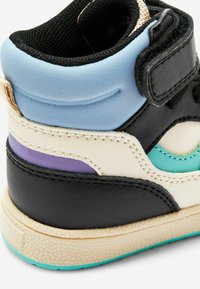 Detailní záběr na multibarevnou špičku vysoce postavené tenisky s černými, krémovými, modrými, fialovými a tyrkysovými panely a texturovanou béžovou podrážkou.