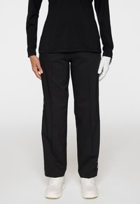 Persoon in zwarte rechte broek, witte sneakers, zwart langmouwshirt en een witte handschoen aan de rechterhand, staande tegen een effen achtergrond.