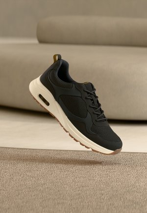 Zapatilla deportiva en blanco y negro con detalles de malla flotando sobre una alfombra beige en un entorno interior minimalista.
