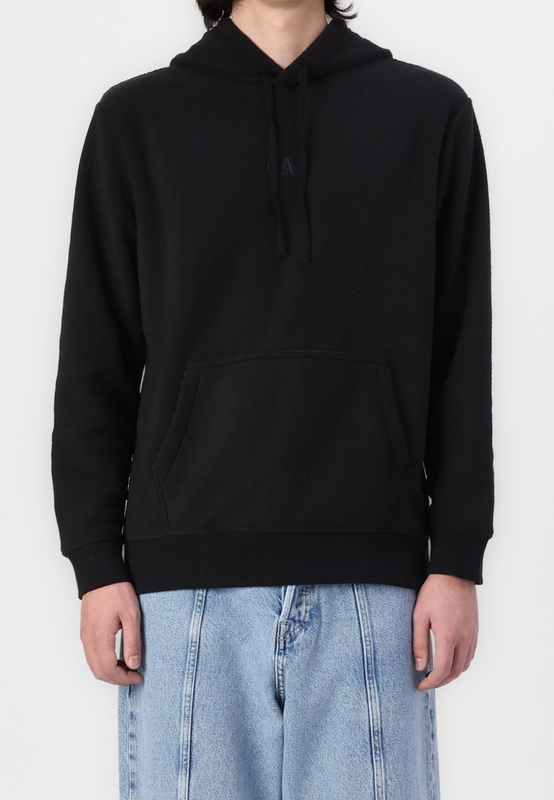 Personne portant un sweat à capuche noir uni avec une poche avant et un jean bleu clair ample sur fond blanc.