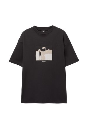 Crna oversized majica s grafičkim zakrpama s bež i bijelim apstraktnim dizajnom. Okrugli izrez i kratke rukave, od pamučnog materijala.