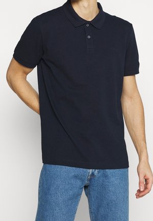 Mand iført sort poloskjorte med korte ærmer og to knapper samt blå jeans, stående foran en hvid baggrund.