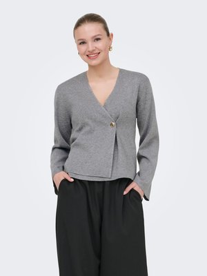 Glimlachende vrouw met blond haar in een grijze wikkeltrui met een gouden knoop, zwarte wijde broek, handen in de zakken.