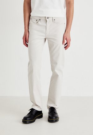 Person trägt gerade geschnittene, hellbeige Jeans und schwarze Schnürschuhe und steht vor einem schlichten weißen Hintergrund.