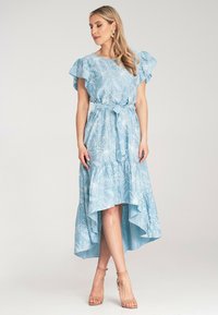 Jasnoniebieska sukienka w kwiatowy wzór, z krótkimi falbanowymi rękawami, paskiem w talii oraz asymetrycznym dołem, wykonana z teksturowanego materiału. Beżowe sandały na obcasie.