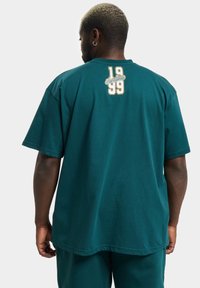 Tyrkysové bavlněné tričko má krátké rukávy, volný střih a na zadní straně logo s nápisem "Rocawear" a čísly "19" a "99".