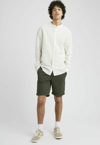 Chemise longue à manches longues de couleur crème claire avec un col montant et un devant à boutons, assortie à un short roulé vert olive et des baskets marron clair.