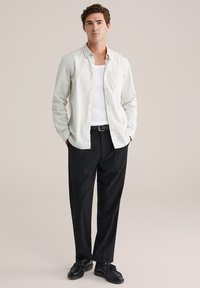 Balta marškinėliai su nupjautomis rankovėmis derinami su juodomis kelnėmis. Marškiniai turi kišenę; bateliai yra juodi, su tekstūruotu paviršiumi ir fringuoto dizaino detale.