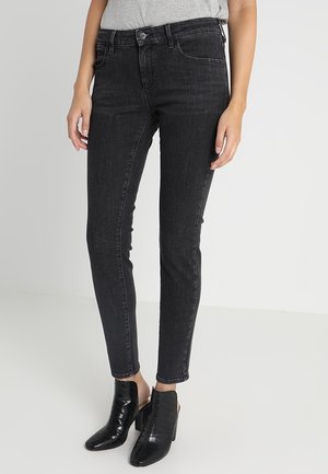 Nainen pukeutuneena mustiin slim fit -farkkuihin ja mustiin korollisiin nilkkureihin, seisoo yksiväristä vaaleanharmaata taustaa vasten.