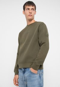 Oliwkowy sweatshirt z okrągłym dekoltem, ściągaczami na rękawach i na dolnej krawędzi. Zawiera subtelne logo na klatce piersiowej oraz naszywkę na rękawie.