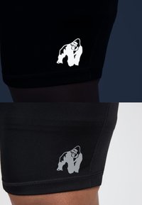 Zwarte sportshorts met een reflecterend gorilla-logo op het linkerpijp. Kenmerkt zich door een gladde textuur en contrasterende stiksels langs de zoom.