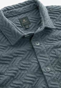 Mittelschichtgraues, strukturiertes Button-Up-Hemd mit umgeschlagenem Kragen und sichtbarem Markenlabel "NEXT" im Nacken.