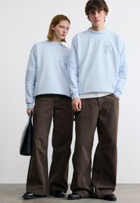 Δύο άτομα φορούν ανοιχτό μπλε φούτερ με την επιγραφή "EST 1978", καφέ φαρδιά παντελόνια και μαύρα παπούτσια· το ένα κρατάει μια μαύρη τσάντα.