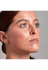 Ritratto ravvicinato di un volto con pelle marrone chiaro, lentiggini visibili, capelli scuri, ciglia lunghe e orecchini a cerchio e a perno. L'illuminazione soffusa mette in evidenza i tratti.