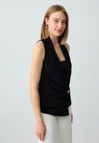 Top nero senza maniche con tessuto drappeggiato, caratterizzato da un accento con bottone dorato. Il tessuto ha una consistenza liscia e una vestibilità comoda. Indossato con pantaloni bianchi.