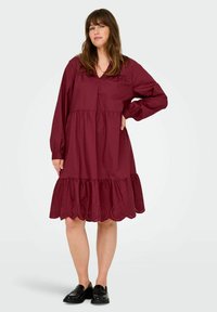 Burgundy cotton dress with long sleeves, V-neck, tiered ruffle hem, and decorative eyelet lace accents at the bottom. Black loafers worn.