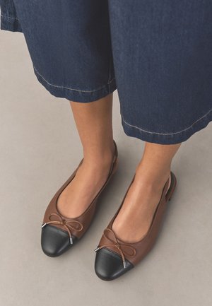 Stopy noszące brązowe i czarne skórzane baleriny z kokardkami, w połączeniu z krótkimi, ciemnoniebieskimi dżinsowymi spodniami.