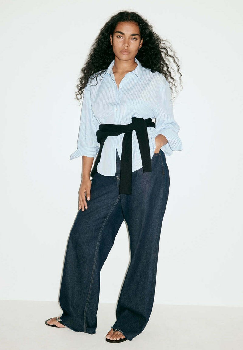 Femme aux longs cheveux bouclés portant une chemise claire à rayures, un jean large foncé, un pull noir noué à la taille et des sandales noires, debout contre un mur blanc.