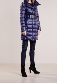 Marineblå pufferjakke med bælte i taljen, høj krave og lynlåsdetaljer, kombineret med mørk denim og sorte ruskindsankelstøvler med høje hæle.