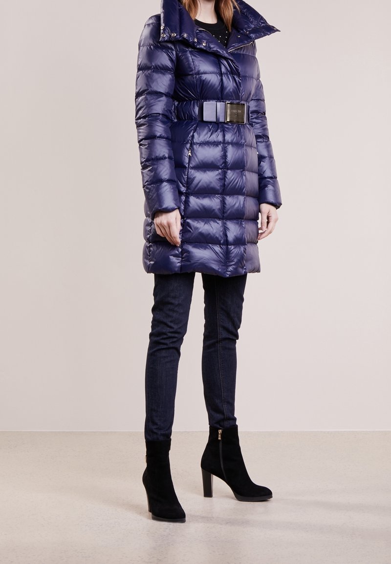 Marineblå pufferjakke med bælte i taljen, høj krave og lynlåsdetaljer, kombineret med mørk denim og sorte ruskindsankelstøvler med høje hæle.