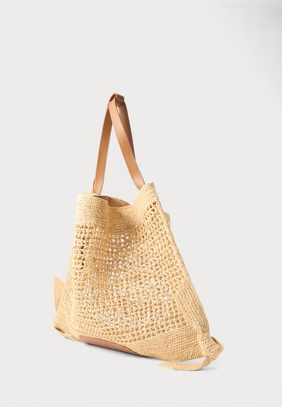 Μπεζ υφασμένη τσάντα ώμου με ανοιχτό πλεκτό σχέδιο και λαδί δερμάτινες λαβές, τοποθετημένη πάνω σε απλό ανοιχτόχρωμο φόντο.