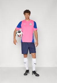 Φανέλα ποδοσφαίρου της Puma με ροζ και μπλε κορυφή, ναυτικές σορτς, λευκές κάλτσες και μαύρα παπούτσια, κρατώντας μία ασπρόμαυρη ποδοσφαιρική μπάλα.