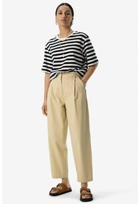 T-shirt rayé noir et blanc, coupe décontractée, associé à un pantalon beige taille haute et des sandales marron avec des accents cloutés.