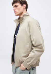 Νέος άνδρας με μπεζ μπουφάν με φερμουάρ και ψηλό γιακά, με τα χέρια εν μέρει στις τσέπες, φοράει σκοτεινό πουκάμισο με λευκές ρίγες, ανοιχτό μπλε τζιν.