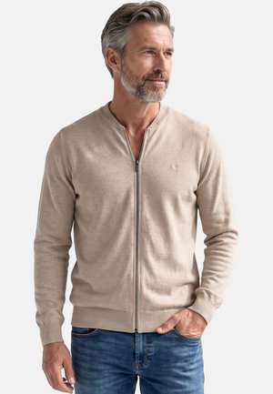 Mann mit grauem Haar und Bart, der einen beigefarbenen Pullover mit Reißverschluss und blaue Jeans trägt, steht mit einer Hand in der Tasche und schaut zur Seite.