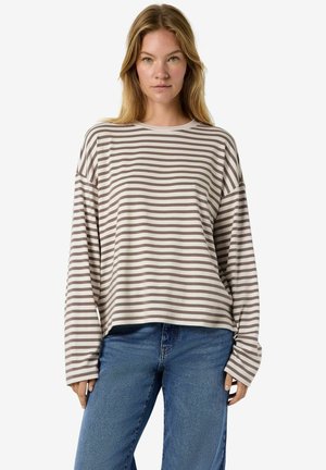 Femme portant une chemise beige et marron rayée à manches longues, ample, et un jean bleu, debout devant un fond clair uni.