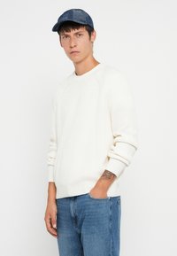 Balta ribota adīta džemperis ar apaļu kaklu, kombinēts ar ziliem džinsiem. Zilā rakstainā cepure papildina apģērbu.