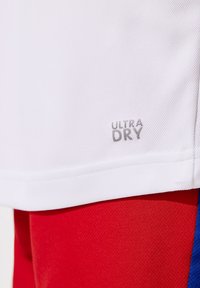 Vit sporttröja med texturerad tyg, försed med "ULTRA DRY" tryckt i silver nära fållen. Bärs med livfulla röda shorts.
