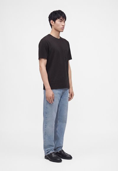Homme debout de profil portant un t-shirt noir à poches, un jean bleu clair et des chaussures en cuir noires sur un fond uni.