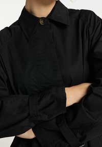 Chaqueta negra de botones con cuello estructurado y mangas largas, que presenta detalles fruncidos en los puños y una textura suave y mate.