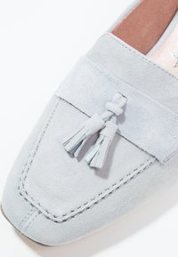 Mocassin en suède gris clair avec une silhouette plate, un bout carré et un accent décoratif de pampilles sur le dessus. Détails de couture subtils partout.