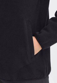 Schwarze Fleecejacke mit seitlicher Tasche, glatter Textur und einem schlichten Design mit rundem Ausschnitt und leicht taillierten Ärmeln.