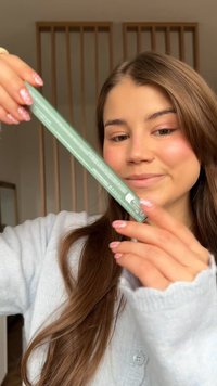 Ung kvinde med langt brunt hår holder og undersøger en slank grøn rektangulær produktæske indendørs iført en lyseblå trøje.